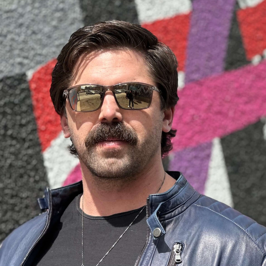 A man with a mustache wearing Murphy shades stands confidently in front of a vibrant graffiti wall.